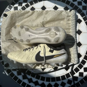Nike Tiempo Legend 10 fotbollsskor ljusbeige - Nike Tiempo Legend fotbollsskor i ljusbeige med svarta detaljer och svart Nike-logga. Ovandel i skinn med textil vid ankeln, snörning och FG-dobbar för spel på gräs men också konstgräs. Snygg design med diskreta guldaccenter och FlyTouch-teknologi för extra komfort. Påse tillkommer också, vid frågor eller funderingar hör gärna av er, pris kan diskuteras! ✅🤩