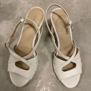 Vita sandaler med klack från Anna Field - Snygga vita sandaler från Anna Field med korsade remmar och öppen tå. Skorna har en elegant hög klack och justerbar rem runt hälen. Tillverkade i syntetmaterial med mjuk innersula för extra komfort. Perfekta för dig som vill ha en stilren och fräsch look.