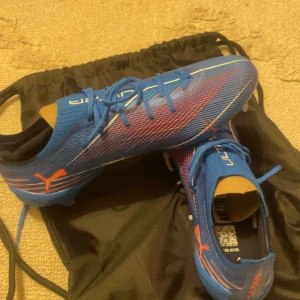 Blåa Puma Ultra fotbollsskor - Snygga blåa Puma Ultra fotbollsskor med röda och vita detaljer. Skorna har snörning, låg profil och är tillverkade i syntetmaterial för lätt känsla och bra grepp på planen. Perfekta för dig som vill sticka ut med stil och fart på fotbollsplanen.