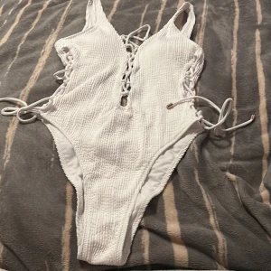 Vit baddräkt med snörning från Censored - Supersnygg vit baddräkt från Censored med ribbad struktur och snörningsdetaljer både fram och i sidorna. Djup urringning och högt skurna ben ger en modern vibe. Perfekt för stranden eller poolen i sommar!