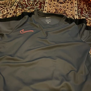 Nike Dri-Fit svart sport t-shirt - Svart Nike Dri-Fit t-shirt med korta ärmar och röd broderad swoosh på bröstet. Tillverkad i lätt och ventilerande polyester, perfekt för träning. Klassisk passform och diskreta svarta paneler på axlarna ger en sportig vibe.