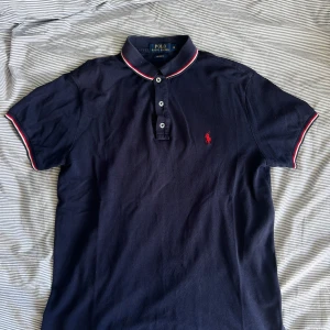 Mörkblå pikétröja från Polo Ralph Lauren - Snygg mörkblå pikétröja från Polo Ralph Lauren i bomull. Tröjan har klassisk krage med röd och vit randig kant, tre knappar framtill och den ikoniska röda loggan broderad på bröstet. Perfekt för en clean och sportig stil.