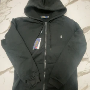 Svart hoodie från Polo Ralph Lauren - Svart hoodie från Polo Ralph Lauren med dragkedja framtill och klassisk huva. Tröjan har vit broderad logga på bröstet och ribbade muddar. Perfekt för en chill och stilren look. Materialet är mjuk bomull som känns skönt mot huden.