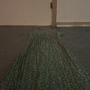 Grön blommig långklänning - Långklänning i ljusgrönt med vitt och rött blommönster. Klänningen har smala axelband och ett luftigt, svepande fall. Perfekt för sommarens varma dagar och har en romantisk vibe. Materialet känns lätt och flowy.