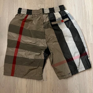 Rutig Burberry shorts - Snygga shorts med klassiskt Burberry-rutigt mönster i beige, svart, vitt och röda detaljer. De har elastisk midja med svart snörning och sidofickor. Perfekta för sommardagar när du vill sticka ut med en ikonisk stil.
