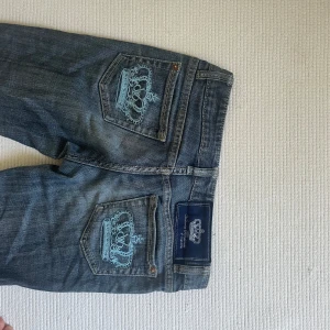 Victoria Beckham blå bootcut jeans - Snygga blå jeans från Victoria Beckham med bootcut passform. Bakfickorna har broderade ljusblå kronor som ger en unik och lyxig vibe. Klassisk femficksmodell i mjukt denimtyg med låg midja och diskret tvättad look.