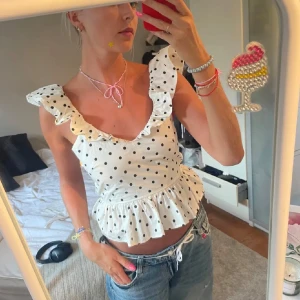 Vit prickig volangtopp med öppen rygg - Supersöt vit topp med svarta prickar, volangdetaljer vid axlarna och v-ringning. Toppen har öppen rygg med knytband och en volangkant nedtill som ger en peplum-effekt. Perfekt för sommarens alla tillfällen!