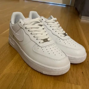 Nike Air Force 1 helvita sneakers - Klassiska Nike Air Force 1 sneakers i helvitt skinn med perforerad tå, vit sula och snörning. Snygg och tidlös modell med låg profil och rund tå, perfekt för dig som vill ha en clean och stilren look. Ikonisk Nike-logga på sidan och detaljer i vitt.