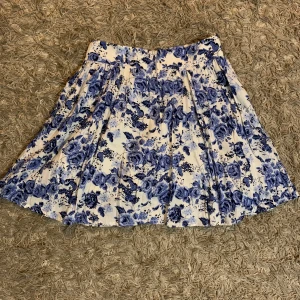 Vit och blå blommig kjol med dragkedja - Superfin kort kjol med blått blommönster på vit botten. Kjolen har veckade detaljer och en dold dragkedja bak för en snygg passform. Perfekt för dig som gillar romantiska vibes och vill sticka ut lite extra i sommar.