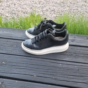 Svarta sneakers från Alexander McQueen - Chunky svarta sneakers från Alexander McQueen med vit, rejäl platåsula och svarta skosnören. Skorna är i skinn och har diskret guldtext på hälen. Perfekta för dig som gillar en clean men edgy stil.