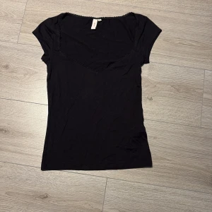 Svart topp med spetskant från Nelly - En svart, figurnära topp från Nelly i storlek XS. Toppen har korta ärmar, v-ringning och en söt spetskant längs halsringningen. Den är använd upp till ungefär 5 gånger!