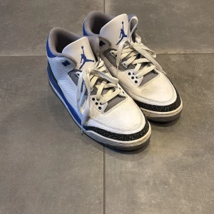 Nike Air Jordan 3 Retro Vit/Blå - Säljer ett par Nike Air Jordan 3 Retro i vit och blå med grå detaljer. Skorna har klassisk snörning, rund tå och ikoniska elefantmönstrade paneler. Ovandelen är i skinn och de har en bekväm vadderad insida. Perfekta för dig som gillar streetstyle och sneakerskultur.