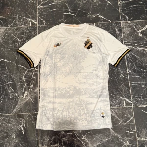 AIK x Nike vit fotbollströja - AIK:s officiella vita fotbollströja från Nike med snyggt stadsmotiv i ljusgrått över hela framsidan. Tröjan har AIK:s klubbmärke på bröstet, gulddetaljer och svarta/gula ränder på ärmsluten. Tillverkad i lätt och ventilerande material, perfekt för match eller träning.