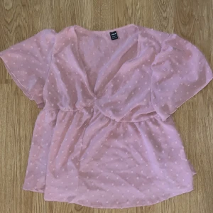 Rosa prickig blus från SHEIN - Söt rosa blus från SHEIN med diskreta prickar, v-ringad hals och luftiga korta ärmar. Modellen har insvängd midja och lätt volang nedtill. Perfekt för dig som gillar en feminin och trendig look.