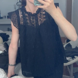 Svart blus med spets och volang - Supersöt svart blus med transparenta prickiga ärmar och detaljerad spets upptill. Volang vid axlarna och lös passform gör den både luftig och trendig. Perfekt att styla med jeans eller kjol för en cool look.