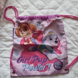Paw Patrol ryggsäck med Skye & Everest - Söt gympapåse/ryggsäck med motiv av Skye och Everest från Paw Patrol. Väskan är lila och rosa med blommor och texten 'Girl Pup Power!'. Praktiska rosa snören och en ögla upptill för enkel upphängning. Perfekt för skola eller träning.