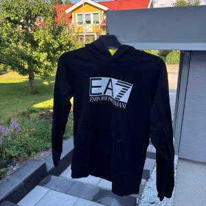 Svart hoodie EA7 Emporio Armani - Svart hoodie från EA7 Emporio Armani med stor vit logga på bröstet. Tröjan har huva, lång ärm och en klassisk känguruficka framtill. Perfekt för dig som gillar stilrena och sportiga plagg med premiumkänsla.