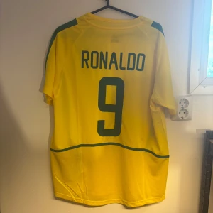 Brasilien Ronaldo 9 fotbollströja Nike - Gul Brasilien fotbollströja från Nike med gröna detaljer, nummer 9 och Ronaldo på ryggen. Klassisk design med korta ärmar, rund hals och officiellt CBF-märke på bröstet. Tillverkad i lätt och ventilerande material, perfekt för fotbollsälskare.