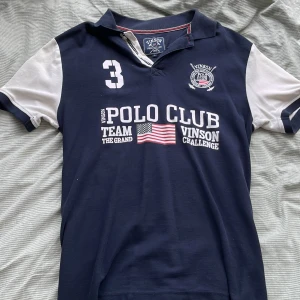 Blå Polo Club t-shirt från Vinson - Mörkblå t-shirt från Vinson Polo Club med vita ärmar och tryck framtill. Klassisk pikékrage, broderad logga och amerikansk flagga på bröstet. Snygg sportig stil med nummerdetalj och kontrastfärgade detaljer.