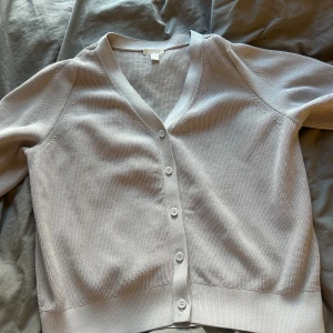 Ljusgrå kofta från COS - En stilren ljusgrå kofta från COS med v-ringning och knappar framtill. Koftan är stickad i bomull och har långa ärmar. Perfekt för lager-på-lager och enkel att matcha med olika outfits.