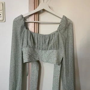Grön omlott blus med långa ärmar - Supersöt ljusgrön blus med små vita blommor, långa ärmar och omlottknytning i midjan. Toppen har rynkade detaljer vid bysten och en fyrkantig urringning som ger en romantisk vibe. Perfekt för vår och sommar!
