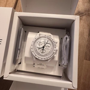 Vit Omega x Swatch MoonSwatch - Säljer en stilren vit Omega x Swatch Speedmaster MoonSwatch. Klockan har en rund boett, kronograf-funktion och tachymeter på ringen. Snyggt vitt textilband med Omega-logga och coolt mån-motiv på baksidan. Perfekt för dig som gillar unik design och ikoniska samarbeten.