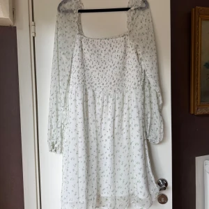 Vit blommig kortklänning med smock vero moda - Supersöt vit kortklänning med små gröna blommor, smockad överdel och långa, transparenta ärmar. Klänningen har fyrkantig ringning och lätt volang i kjolen. Använd 1 gång.