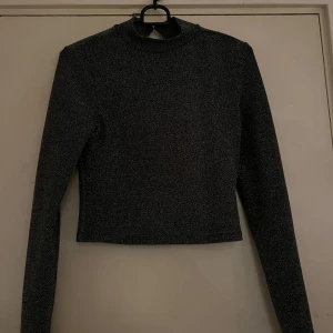 Glittrig svart croppad polotopp - Snygg svart glittrig polotopp med lång ärm och croppad passform. Toppen har en öppen rygg med en cool cut out-detalj och hög hals. Perfekt för dig som vill sticka ut med en trendig look och gillar att glänsa.