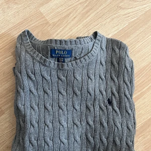 Grå kabelstickad tröja Polo Ralph Lauren - Kabelstickad grå tröja från Polo Ralph Lauren i storlek XL. Klassisk rund halsringning, ribbade muddar och broderad logga på bröstet. 
