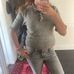 Grå stickad tröja med volangdetalj - Superfin grå stickad tröja med korta ärmar och volangdetaljer vid halsringningen och ärmsluten. Tröjan har en söt knappslå framtill och är figurnära i modellen. Perfekt att styla med jeans för en chill look.