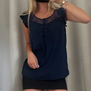 Mörkblå blus med spets och knappar - Storlek XS från Vero Moda💞Supersöt mörkblå blus med transparent spets upptill och dekorativa små knappar framtill. Blusen har ärmlös design med lätt volang vid axlarna och rund halsringning. 