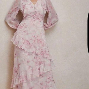 Lång blommig volangklänning i rosa - Helt ny använd 1 gång Romantisk långklänning med rosa blommigt mönster på ljus botten. Klänningen har v-ringning, knappar framtill, långa puffärmar och flera volangdetaljer. Perfekt för sommarens alla tillfällen. Materialet är lätt och luftigt, troligtvis polyester eller liknande.