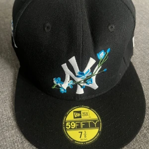 New Era Yankees keps med blommor - Snygg svart New Era 59FIFTY keps med New York Yankees-logga framtill, dekorerad med broderade blå blommor. På sidan finns ett broderat World Series-märke i blått och vitt. Klassisk rak skärm och broderad New Era-logga på sidan.