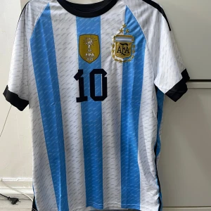 Argentina Messi 10 fotbollströja - Säljer en officiell Argentina fotbollströja med Messi och nummer 10. Tröjan har klassiska blå och vita ränder, broderat AFA-märke med tre stjärnor och FIFA 2022-märke. Svarta detaljer vid krage och ärmslut. Tillverkad i lätt och ventilerande material, perfekt för match eller träning.