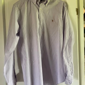 Ljuslila skjorta från Ralph Lauren - Säljer en klassisk ljuslila skjorta från Ralph Lauren i 100% bomull. Skjortan har lång ärm, knappar framtill och den ikoniska broderade loggan på bröstet. Perfekt för dig som gillar stilrena och tidlösa plagg. En tråkig fläck på framsidan, skriv om ni vill veta mer.
