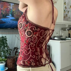  korsett med snörning - Säljer en bordeauxröd korsett med vackra broderade blommor och swirl-detaljer i guld.  Toppen har halterneck, öppen rygg med snörning och croppad passform. 