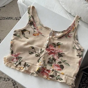 Blommig croppad linnetopp med volang - Croppad linnetopp från HM Divided i beige med blommigt mönster i rosa, grönt och gult. Toppen har ribbad struktur, knappar framtill och volangkant nedtill. 