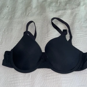 Svart vadderad bh med bygel - En klassisk svart bh med vadderade kupor och bygel för extra stöd. Justerbara axelband och hakknäppning bak. Slät och enkel design som funkar under det mesta. Mjukt och stretchigt material för bekväm passform.Tror ca 70A eller xxs.