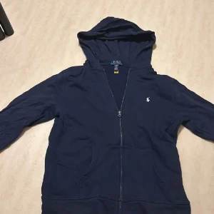 Mörkblå hoodie från Polo Ralph Lauren - Säljer en mörkblå hoodie från Polo Ralph Lauren i storlek barnstorlek XL. Tröjan har dragkedja, huva och känguruficka framtill. Klassisk broderad logga på bröstet. Perfekt för dig som gillar stilren och bekväm streetwear.