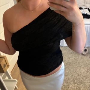 Svart oneshoulder topp i spets - Säljer en svart oneshoulder topp med snyggt spetsmönster och croppad passform. Toppens struktur är lätt transparent och ger en cool vibe. Perfekt för dig som vill sticka ut med en asymmetrisk look.