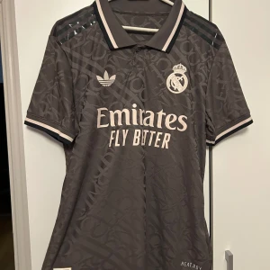 Real Madrid Vini Jr. matchtröja Adidas - Snygg mörkgrå Real Madrid fotbollströja från Adidas med Vini Jr. och nummer 7 på ryggen. Tröjan har korta ärmar, klassisk krage och diskret mönster i tyget. Broderad klubbmärke och Adidas-logga framtill, samt Emirates Fly Better-tryck. Tillverkad i lätt funktionsmaterial.