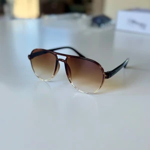 Bruna aviator-solglasögon med tonade glas - Snygga aviator-inspirerade solglasögon med bruna bågar och tonade glas som går från mörkbrunt upptill till ljust nedtill. Klassisk form med svarta skalmar och metall-look. Perfekta för att lyfta din stil under soliga dagar.