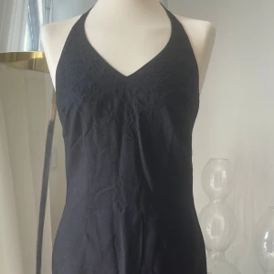 Svart halterneck klänning med broderi - Svart kortklänning med halterneck och v-ringning. Överdelen har broderade blommor som ger en snygg detalj. Klänningen har dragkedja i ryggen och är figurnära i modellen. Perfekt för dig som gillar stilrena och feminina plagg. I finaste kvalitet. 