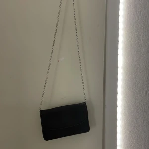 Svart kuvertväska med kedja - Snygg svart kuvertväska med silverfärgad kedja som axelrem. Väskan har ett stilrent ormskinnsmönster och stängs med magnetknapp. Perfekt storlek för det viktigaste och ger en cool touch till din outfit.