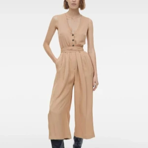 Beige byxdress med knyt och knappar - Snygg beige byxdress med vida ben och croppad längd. Djup v-ringning, knappar framtill och knytband i midjan. Tillverkad i ett mjukt, lätt material som känns luftigt. Perfekt för dig som gillar en avslappnad men trendig look.