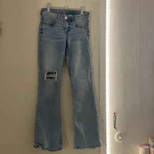 Ljusblå bootcut jeans med slitning från H&M - Säljer ett par ljusblå low waist bootcut jeans med snygg slitning och hål på ena knät. Jeansen har klassisk femficksdesign och är tillverkade i mjukt denimtyg. Perfekta för dig som gillar en avslappnad och trendig look. OBS använda en gång.