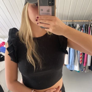 Svart croppad topp med volangärmar - Säljer en svart croppad topp med volangdetaljer på ärmarna. Toppen har en tight passform och är tillverkad i ett ribbat material som ger en snygg struktur. Liten i stl passar både S och M 