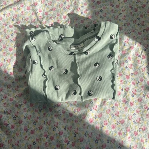 Ljusgrön långärmad topp med yin yang-mönster  - Superfin ljusgrön långärmad topp från Kappahl med svarta och vita yin yang-symboler över hela. Toppen har ribbad struktur och vågiga volangdetaljer längs sömmarna framtill. Perfekt för dig som gillar unika detaljer!