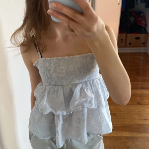 Ljusblå volangtopp från Gina Tricot - Supersöt ljusblå topp från Gina Tricot med smock upptill och volanger nedtill. Toppen är ärmlös och har en luftig och somrig känsla, perfekt för varma dagar. Materialet är mjukt och lätt, vilket gör den extra bekväm.