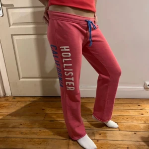 Rosa mjukisbyxor från Hollister - Rosa mjukisbyxor från Hollister med blå dragsko i midjan och tryckt text längs benet. Byxorna har en avslappnad passform och är perfekta för chill dagar hemma.